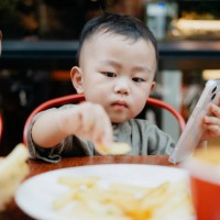 asian toddler boy eating french fries and playing on smartphone in a restaurant - junk food stock pictures, royalty-free photos & images