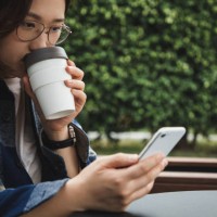 asian woman enjoying coffee using reusable cup while using her phone - junk food stock pictures, royalty-free photos & images