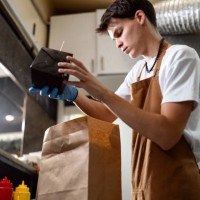 at the fast food restaurant kitchen, male teenager packing the order into brown paper bag - junk food stock pictures, royalty-free photos & images