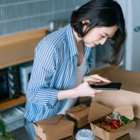 beautiful and cheerful young asian woman standing by the kitchen counter, taking a photo of delicious home delivery takeaway fresh healthy salad lunch box with smartphone, sharing on social media before eating it. eating at h