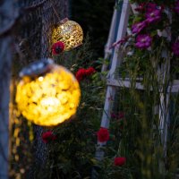beautiful round garden lanterns with carved floral patterns hang on a fence in an evening garden among flowering plants and glow with yellow light - garden decoration stock pictures, royalty-free photos & images
