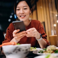 beautiful smiling young asian woman taking photos of scrumptious traditional thai food served on dining table with smartphone while enjoying lunch in a thai restaurant. eating out lifestyle. camera eats first culture - food s