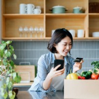 belle jeune femme asiatique souriante faisant ses courses en ligne avec l’appareil d’application mobile sur le smartphone et faisant le paiement en ligne avec sa carte de crédit, avec une boîte d’épicerie organique colorée et