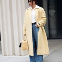 Benthe Liem wears blue jeans, white shirt, pale yellow long coat, yellow bag with attached black mini bag outside the Herskind fashion show during...