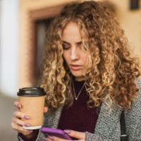blonde curly girl with paper cup and mobile phone in hand in coat outdoors - junk food stock pictures, royalty-free photos & images