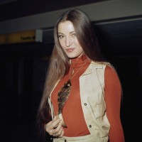 British actress Jane Seymour, wearing a corduroy waistcoat over a red turtleneck sweater, holding the tail of the large gold fish pendant hanging...