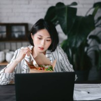 busy young asian woman eating takeaway healthy salad lunch box while working from home using laptop and handling paperworks in the living room at home. remote working concept - junk food stock pictures, royalty-free photos & 