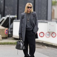Camille Charriere wears gold necklaces, black sunglasses,, a grey sweater, a grey coat, black trousers, a grey silver bag and beige sneakers outside...