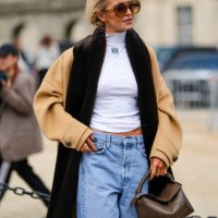 Caroline Daur wears dark brown sunglasses, white turtleneck Loewe top, light brown dark brown Loewe coat, light blue denim jean pants, dark green...