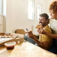 cheerful couple using cell phone and eating pizza on a break from home renovation process. - junk food stock pictures, royalty-free photos & images