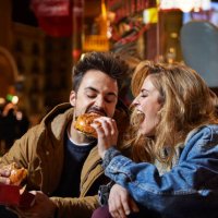 cheerful woman feeding burger to man at night - junk food stock pictures, royalty-free photos & images