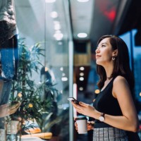 cheerful woman holding a cup of coffee, checking her smartphone while standing outside a boutique looking at shop window in the evening in the city - fashion stock pictures, royalty-free photos & images