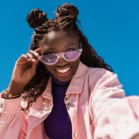 cheerful woman with braided hair lowering sunglasses, revealing bright smile beneath cloudless azure background - fashion stock pictures, royalty-free photos & images
