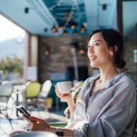 cheerful young asian woman using smartphone while sitting at a sidewalk cafe alone during free time, enjoying time on her own on a beautiful sunny day. lifestyle and technology. slow living. sustainable lifestyle - travel sto