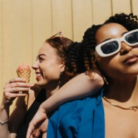 cheerful young woman eating ice cream while leaning on fashionable female friend against corrugated wall - fashion stock pictures, royalty-free photos & images