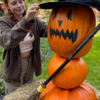 close-up image of attractive, red-haired woman arranging a witch's hat on pumpkin witch, three orange gourds stacked to make 'witch' with broomstick, surrounded by hay, halloween face jack o'lantern outdoors on lawn, focus on