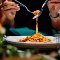 close-up, mid-section of young asian woman sitting at dining table holding cutleries, twirling freshly served spaghetti bolognese and enjoying her meal in restaurant. italian food. dining out. people, food and lifestyle - foo