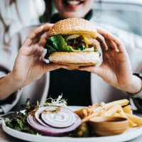 close up of happy young asian woman sitting by the window in a restaurant, enjoying lunch during the day. she is having freshly made delicious burger with fries and side salad. lifestyle, people and food concept - junk food s