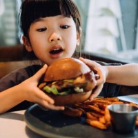 close up shot of funny little asian girl looking excited while holding a big burger and making a face while enjoying eating burger with sweet potato fries in a restaurant. eating out lifestyle - junk food stock pictures, roya