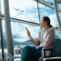 confident young asian businesswoman with suitcase looking ahead while using smartphone waiting for her flight at airport terminal. business traveller. woman in business. business lifestyle and technology. travel and vacation 