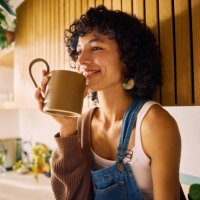 content woman savoring the aroma of her morning coffee in a well-lit kitchen with modern wooden accents and lush houseplants, embracing a moment of serenity, symbolizing mindfulness and the joy of simple pleasures in everyday