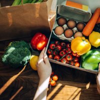 cropped hands of woman opening a home delivery cardboard box filled with multi-coloured fresh organic fruits, vegetables and groceries on wooden background against sunlight. grocery subscription boxes. meal kits. convenient a