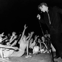 Crowd of screaming teenage girls reach for Elvis Presley as he sings on stage.