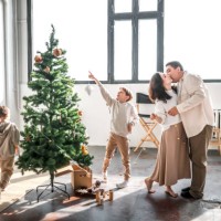 cute family photo of preparations for christmas: middle-aged parents kiss, and their children sons run around christmas tree, pulling on festive garland - home decoration stock pictures, royalty-free photos & images
