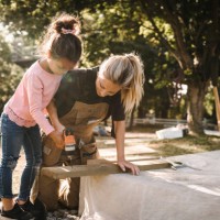 daughter using drill machine on wooden plank by mother in backyard - home decoration stockfoto's en -beelden