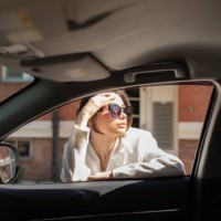 delightful woman standing near automobile, leaning on open window, softly touching forehead and looking away through classy sunglasses, posing on backdrop of city buildings. - fashion stock pictures, royalty-free photos & ima