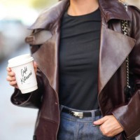 Diane Batoukina wears a black t-shirt, a dark brown shiny leather long coat, a black shiny grained leather Kelly small belt from Hermes, black denim...