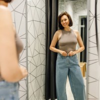 elegant woman with brown hair looking at herself in mirror trying on new denim jeans and beige top in fitting room - fashion fotografías e imágenes de stock