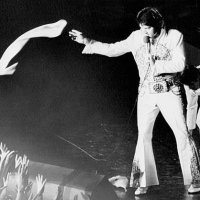 Elvis Presley, in a white jumpsuit with gold brocade and studs, tosses a nylon scarf into the waiting hands of the fans at a concert in the...