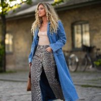 Emili Sindlev wears a blue denim long coat, a beige tank-top from Miu Miu, beige and black leopard print pattern denim flared pants, a camel shiny...