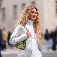 Emili Sindlev wears earrings, a silver shiny jacket, a white turtleneck top, a green leather Coach bag, white pants with printed green apples,...