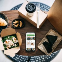 flat lay of takeaway food ordering mobile app on smartphone, with assorted takeaway dishes, healthy grilled chicken vegetable salad, croissant, japanese style rice ball / onigiri and a cup of coffee freshly served on coffee t