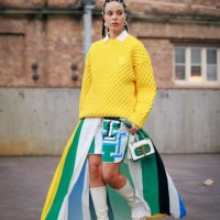 Fleur Egan wearing buttons on her hair, yellow jumper, green and blue mixed colour maxi split skirt, white boots and green and white bag during...