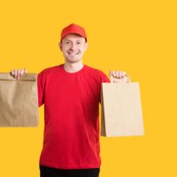 food delivery service from an online store. courier in red uniform with craft bags gives fast food order on yellow isolated background. copy space - junk food stock pictures, royalty-free photos & images