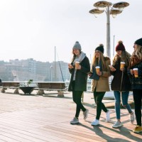 four friends with coffee to go and cell phones walking on promenade in winter - junk food stock pictures, royalty-free photos & images