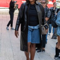 Gabriella Karefa-Johnson is seen wearing a brown leather coat, blue top, blue denim shorts, black sunglasses, black shoes and carrying a grey bag...