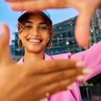 gen z woman framing her face with her hands and smiling. close-up shot with modern building in the background. positive emotions and urban lifestyle concept. - fashion stock pictures, royalty-free photos & images