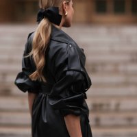 Ginta is seen wearing a black bow around her ponytail, gold drop-shaped earrings, a round diamond stud earring, an oversized black leather jacket...