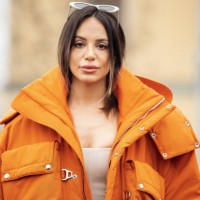 Guest is seen wearing white sunglasses, an orange oversized cargo parka with silver details, a white corset, white trousers, and orange velvet...