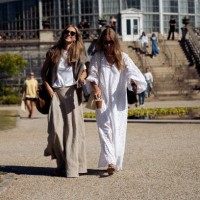 Guest wearing long grey skirt, white T-shirt, brown sweater over the shoulders, cup of ice coffee and big brown bag and a guest wearing long white...