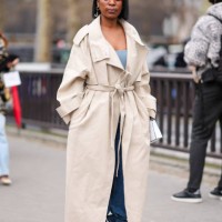 Guest wears a beige trench coat, a blue low neck top, a blue denim jeans / pants, black leather high heels shoes with golden chains, outside Chanel,...