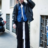 Guest wears a black and white printed bandana, a white lace as necklace, a black shirt, a denim gilet, a black leather jacket and black wide leg...