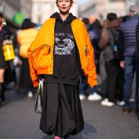 Guest wears a black hoodie sweater, a neon orange bomber jacket, a black oversized pleated skirt, silver shiny boots, a silver shiny bag, outside...