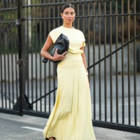 Guest wears a black leather bag, earrings, a yellow sleeveless top, a pale pastel yellow pleated long skirt, sandals, outside COS, during New York...