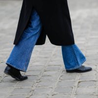 Guest wears a black long coat, blue denim flared pants, black shiny leather ankle shoes, outside Chanel, during Paris Fashion Week - Haute Couture...