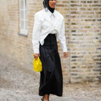 Guest wears a black scarf as a headband, a silver chain hat, a white asymmetric cropped blazer jacket, a white shirt, a black high waist long skirt,...